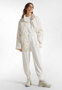 Gepolsterte cremefarbene Jacke mit Druckknopfverschluss, kombiniert mit einem weißen Hoodie und lockeren Jogginghosen. Beigefarbene Sneaker mit grünen Akzenten.