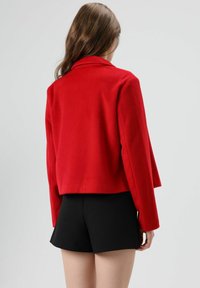 Rode, cropped blazer van wol met een reverskraag en lange mouwen. Gecombineerd met zwarte shorts. Glad oppervlak, minimalistisch ontwerp.