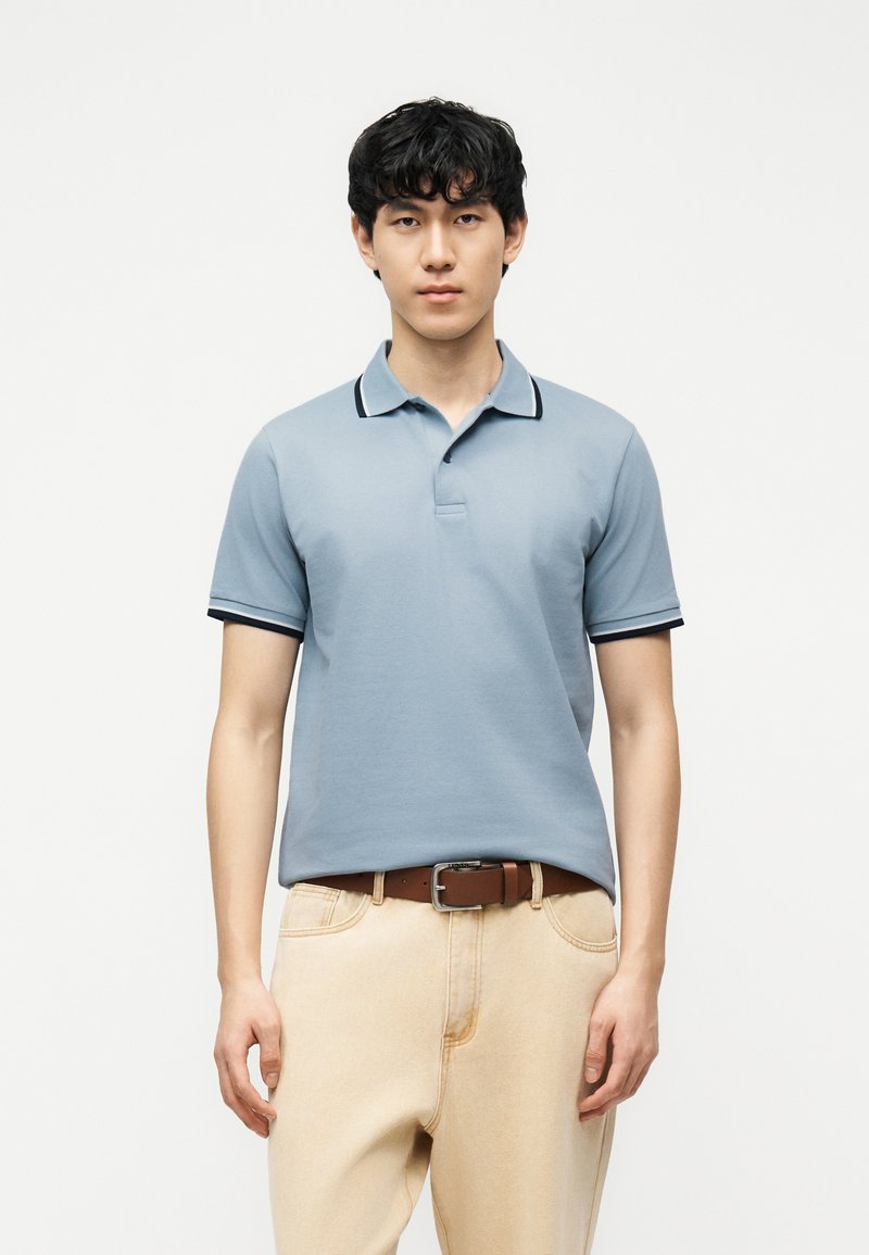 Νεαρός με μαύρα κατσαρά μαλλιά, που φοράει ανοιχτό μπλε πόλο πουκάμισο φορεμένο μέσα σε μπεζ παντελόνι με καφέ ζώνη, στέκεται μπροστά σε απλό λευκό φόντο.