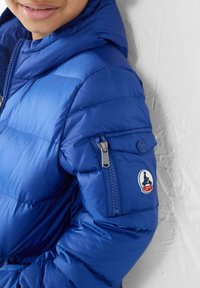 Veste matelassée bleue avec capuche, caractérisée par des sections matelassées et une poche zippée sur le bras. Comprend un patch de marque sur la manche.