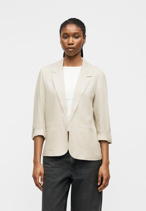 Vrouw met gevlochten haar die een beige blazer met opgerolde mouwen draagt, een wit topje en donkergrijze jeans, staand tegen een effen lichte achtergrond.