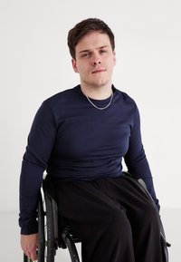 Sötétkék, hosszú ujjú ing fekete nadrággal párosítva, egy kerekesszékben ülő személyen, ezüst láncnyaklánc mellett.