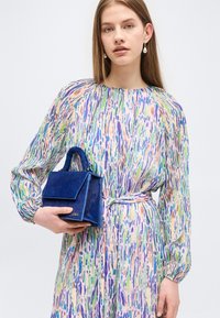 Jeune femme portant une robe multicolore à manches longues et tenant un petit sac à main bleu avec une anse supérieure, sur un fond clair.