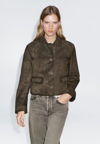 Brown suede cropped jacket with a pointed collar, front buttons, and two side pockets, worn over grey jeans against a plain background.