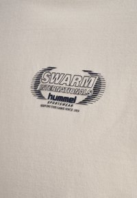 Logo SWARM INTERNATIONALS z stylizowanymi liniami, marka odzieży sportowej HUMMEL oraz napis „SŁUŻYMY GRZE OD 1923” na beżowej tkaninie.