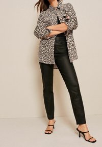 Camicia in stampa leopardata beige e nera, abbinata a pantaloni neri simili alla pelle e tacchi a spillo neri. La camicia presenta dettagli con bottoni e maniche arrotolate.