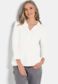 Camicia bianca a maniche lunghe con scollatura a V, patta con bottoni e pieghe verticali. Tessuto morbido in una vestibilità comoda, abbinata a pantaloni grigi.