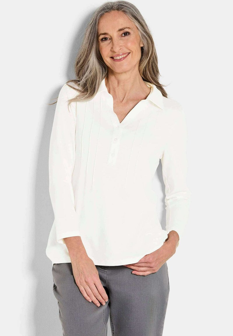 Camicia bianca a maniche lunghe con scollatura a V, patta con bottoni e pieghe verticali. Tessuto morbido in una vestibilità comoda, abbinata a pantaloni grigi.