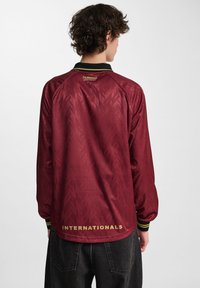 Fiatal, göndör hajú személy bordó hosszú ujjú sportos felsőt visel, rajta a "hummel" logóval és hátul az "INTERNATIONALS" felirattal.