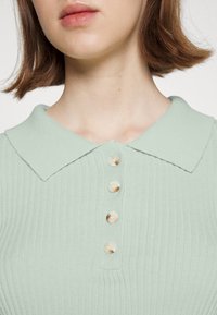 Camisa polo de malha verde clara com colarinho e cinco botões bege na parte da frente; a textura ribada é proeminente em toda a camisa.