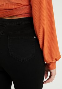 Jeans negros de tiro alto con un bolsillo trasero que presenta un pequeño adorno plateado, combinados con una camiseta de manga larga acanalada naranja.