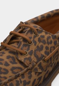 Chaussure bateau en cuir marron avec imprimé léopard foncé et lacets en cuir beige passés à travers des œillets en métal.