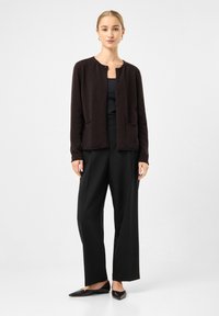 Brauner Cardigan mit Rundhalsausschnitt, Knopfverschluss und zwei vorderen Taschen, kombiniert mit schwarzen weit geschnittenen Hosen und spitzen Ballerinas.