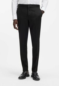 Pantaloni neri su misura realizzati in tessuto liscio, con fronte piatto, pieghe e tasche laterali. Abbinati a scarpe di pelle scura.