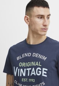 Marineblå bomulls T-skjorte med vintage grafikk i hvitt og grønt, med rund hals og korte ermer. Glatt tekstur, avslappet passform.