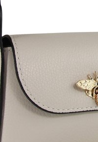 Beige leren handtas met een gestructureerde afwerking, voorzien van een klep-sluiting en een gouden accent in de vorm van een bij. De randen zijn afgewerkt in zwart.