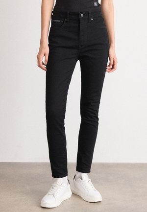 Jeans skinny pretos com uma textura suave, cintura alta, bolsos frontais e cinco passantes para cinto. Combinados com ténis brancos.