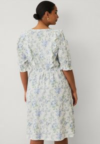 Robe fleurie de couleur claire avec des motifs bleus et verts. Présente des manches volantées et une taille smockée. Mi-longue, fabriquée en tissu texturé.