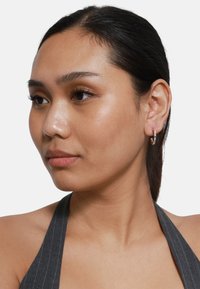 Orecchino a cerchio argentato, finitura liscia, leggermente riflettente. Il modello indossa un top halter a righe grigie, mettendo in evidenza chiaramente il design dell'orecchino.