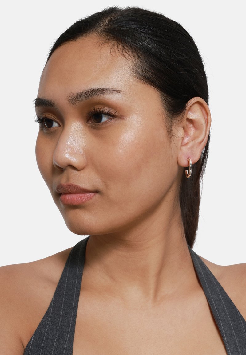 Orecchino a cerchio argentato, finitura liscia, leggermente riflettente. Il modello indossa un top halter a righe grigie, mettendo in evidenza chiaramente il design dell'orecchino.