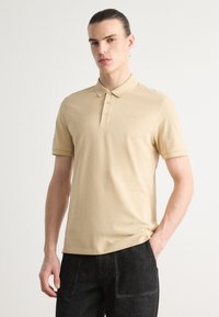 Beige színű pólóing klasszikus gallérral, három gombos megoldással és hímzett logóval a mellrészen. Rövid ujjak és puha szövettextúra.