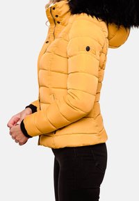 Giacca a vento gialla con cappuccio foderato in pelliccia, caratterizzata da un design trapuntato, chiusura frontale con zip e polsini in maglia nera. Vista laterale.