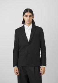Black blazer with notched lapels, single button closure, two front pockets, and a smooth texture, paired with a white button-up shirt.