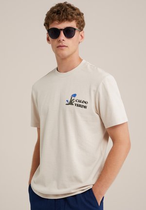 Beige cotton t-shirt with short sleeves, featuring a blue floral design and the text "CALDO VERDE" in black. Worn with dark sunglasses.