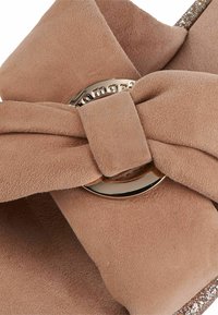 Scarpa in suede beige con dettagli di cinturino annodato, caratterizzata da un accento metallico rotondo e un tacco lucido e scintillante. Texture complessivamente liscia.