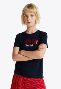 T-shirt in cotone navy con grafica rossa "TOMMY New York", maniche corte, scollo rotondo, indossato con una gonna rossa con pois bianchi.