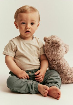 Lysbeige skjorte med korte ærmer, en lomme og knapdetaljer, parret med grønne bukser med opklappede ben. En blød, lysebrun teddy bjørn sidder ved siden af barnet.