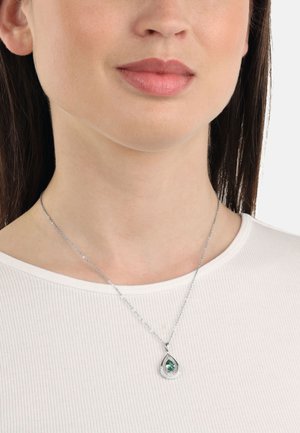 Collana d'argento con un pendente a goccia che presenta una pietra verde e decorazioni, appoggiata su un top bianco a righe.