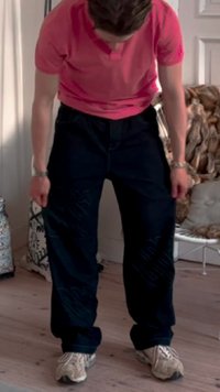 Dark blue baggy pants with a textured fabric. Worn with a short-sleeve pink top and white sneakers. Standing on a wooden floor.