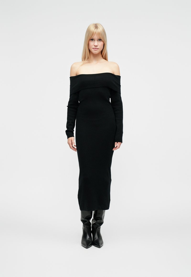 Vestito nero in maglia scollato sulle spalle con maniche lunghe, silhouette aderente, lunghezza fino a metà polpaccio, abbinato a stivali neri al ginocchio.