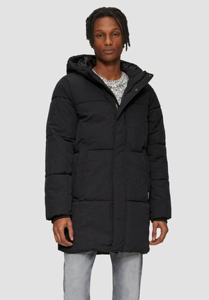 Manteau long noir rembourré avec capuche, doté d'une fermeture éclair à l'avant, de poches latérales et d'une texture matelassée. Porté par-dessus un pull léger à motifs.