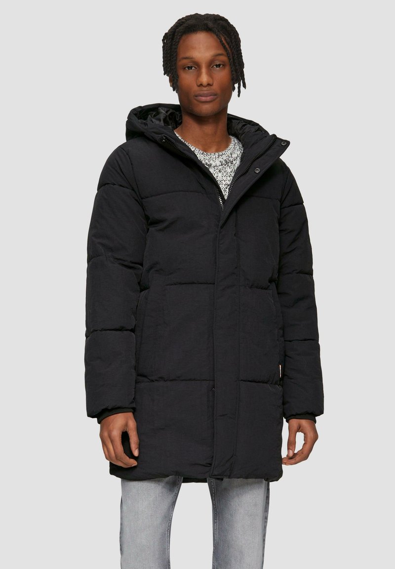 Padded schwarzer langer Mantel mit Kapuze, mit Reißverschluss vorne, seitlichen Taschen und gesteppter Struktur. Trägt man über einem hellen, gemusterten Pullover.