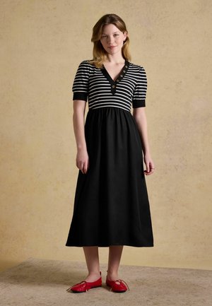 Femme debout portant une robe midi noire avec un haut rayé noir et blanc et des boutons, associée à des ballerines rouges, fond beige texturé.