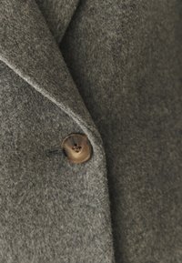 Abrigo de lana gris con una tela suave y texturada, con un gran botón marrón brillante en el cuello. Las solapas entalladas realzan el diseño.