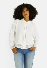 Camisa blanca de botones con una ribete de encaje en la cintura y los puños, tejido texturizado, cuello clásico y un ajuste relajado. Se lleva con jeans azules.