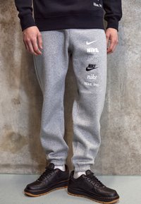 Grå sweatpants med olika Nike-logotyper i vitt och svart. Har elastiska muddar och en avslappnad passform, tillsammans med svarta Nike-skor.