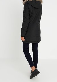 Svart parka med fuskpäls på huvan, långa ärmar och rak skärning, kombinerad med mörka leggings och svarta skor.