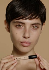 Correttore KIKO Skin Tone in un tubo beige con tappo nero, tenuto da una persona con capelli corti contro uno sfondo marrone chiaro uniforme.