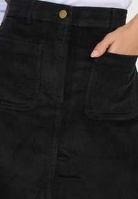 Mustahdud hitam dengan tekstur ribbed vertikal, dua saku depan, dan penutupan tombol kuningan di pinggang.