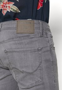 Tylny widok na szare dżinsy z denimu z skórzaną naszywką marki Jack & Jones oraz włożoną w nie kwiatową koszulę.