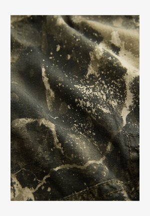 Zaļa auduma teksturēts virsmas dizains ar raibu bēša rakstu. Tuvplāns atklāj smalkas detaļas un precīzu šuvumu.