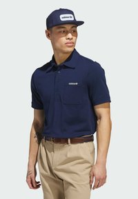Mørkeblå polo-skjorte med knappedesign og brystlomme, med en hvit adidas-logo. Kombinert med beige bukser og en marinblå caps.