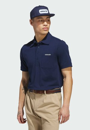 Polo shirt bleu marine avec un design à boutons et une poche poitrine, arborant un logo adidas blanc. Associé à un pantalon beige et une casquette marine.