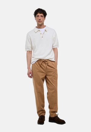 Homme portant un polo blanc à manches courtes, un pantalon beige avec cordon de serrage et des chaussures marron foncé à lacets, debout devant un fond uni.