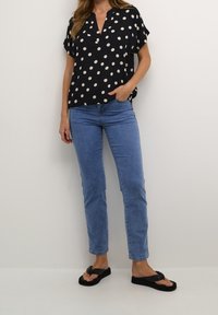 Mørkeblå kortærmet bluse med store hvide prikker, der har V-udskæring og en afslappet pasform, parret med lyseblå lige ben jeans og sorte sandaler.