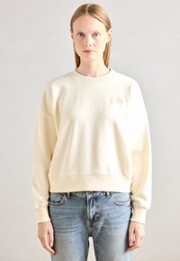 Sweatshirt crème avec un col et des poignets côtelés. Présente une coupe courte et un logo brodé subtil sur la poitrine. Assorti avec un jean bleu clair.
